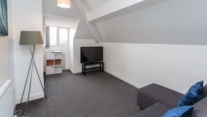A modern living room with a grey sofa, a flat-screen TV on a stand, a floor lamp, and a window with curtains. - Cherry Property - Blueberry Suite (Blackpool)
