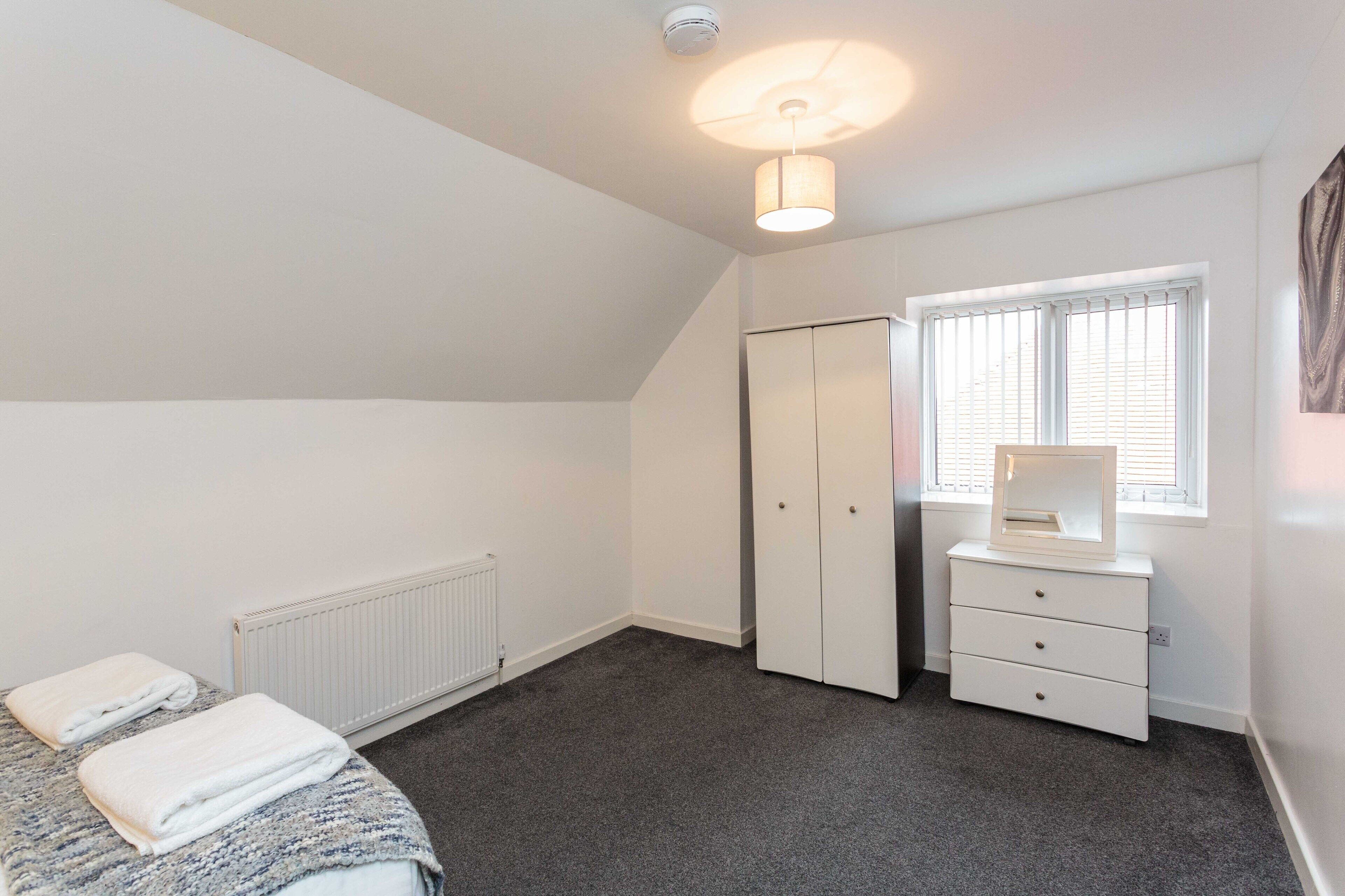 A bedroom with a bed, a dresser, a radiator, a window with blinds, and a ceiling light.