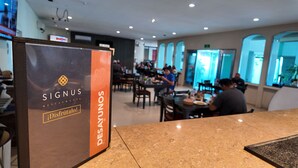 Interior de un restaurante con un cartel que dice 'SIGNUS RESTAURANTE' y un cartel que indica 'DESAYUNOS'. Las mesas están dispuestas y hay comensales.