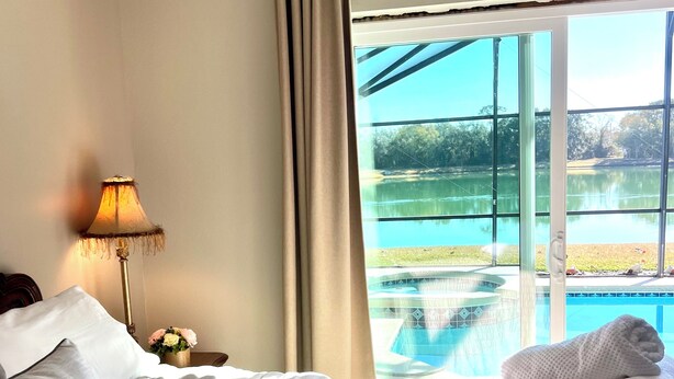 A hotel room with a bed, a lamp, a nightstand with flowers, and a view of a swimming pool and lake. - MAGIC 5* LAKE VIEW VILLA HEATED POOL&HOT SPA, near DISNEY, 10 Guests (Kissimmee)