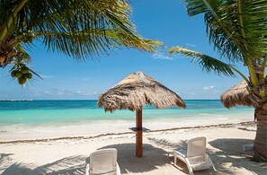 A beach with white lounge chairs, a thatched umbrella, and palm trees.