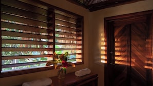 A room with wooden shutters, a window with a view of greenery, a wooden door, and a small table with towels and a vase. - Totoco Eco Resort (Altagracia)