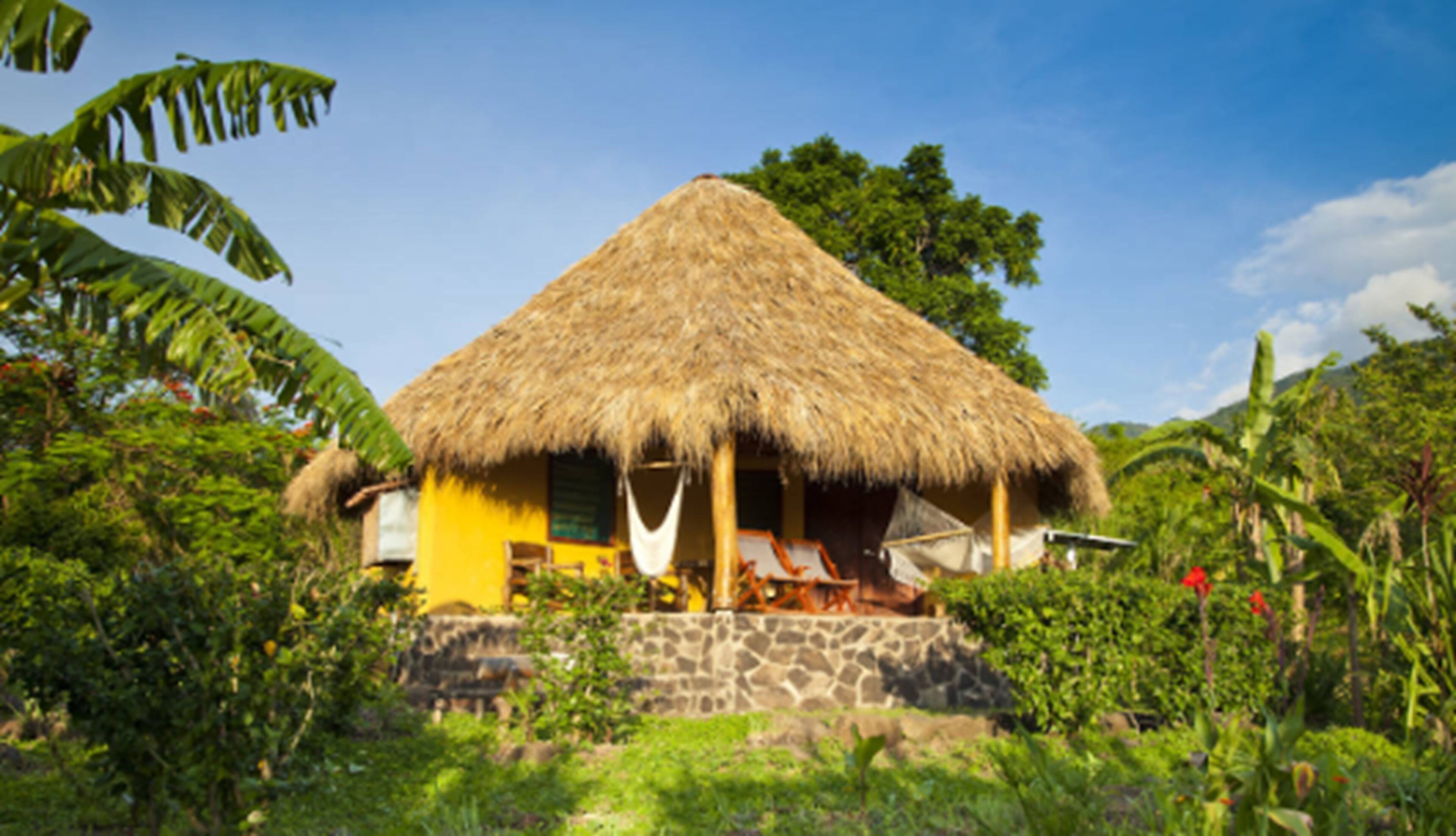 A thatched-roof house with a stone foundation, surrounded by lush greenery and a clear sky.