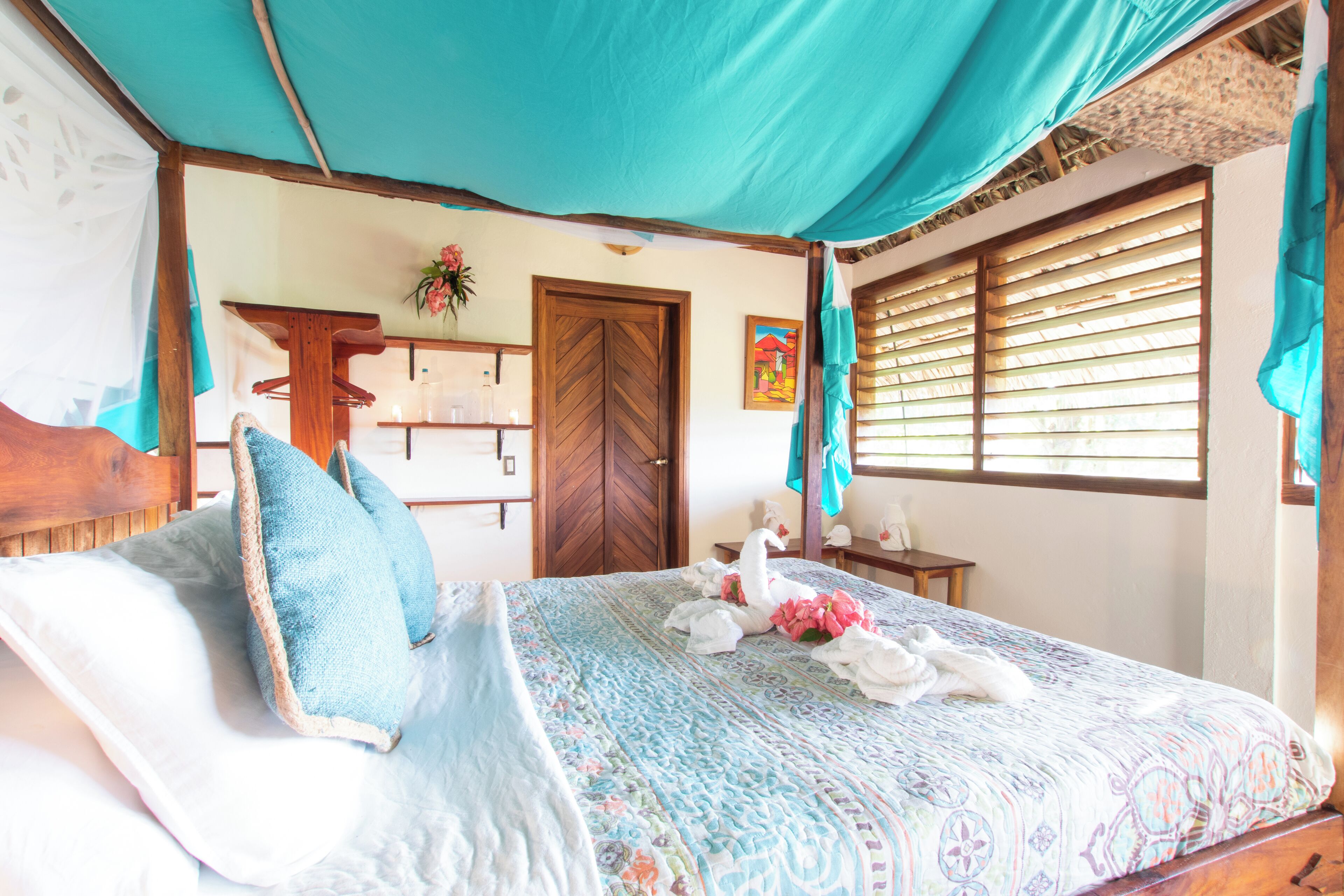 A bedroom with a bed, a wooden door, a window with blinds, and a small table with a vase.