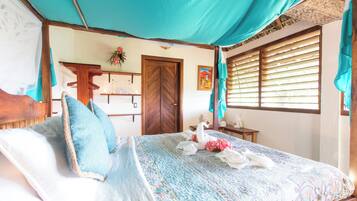 Un dormitorio con cama, puerta de madera, ventana con persianas y una pequeña mesa con un florero.