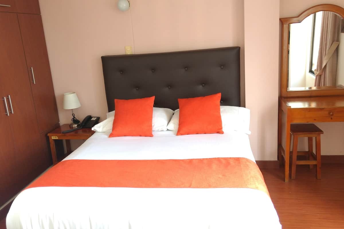 Una cama cuidadosamente hecha, con un cabecero oscuro, dos almohadas naranjas y una colcha blanca. Junto a la cama, una mesita de noche de madera con una lámpara y un teléfono. Un escritorio de madera y una silla están situados al lado de un espejo grande en la pared.