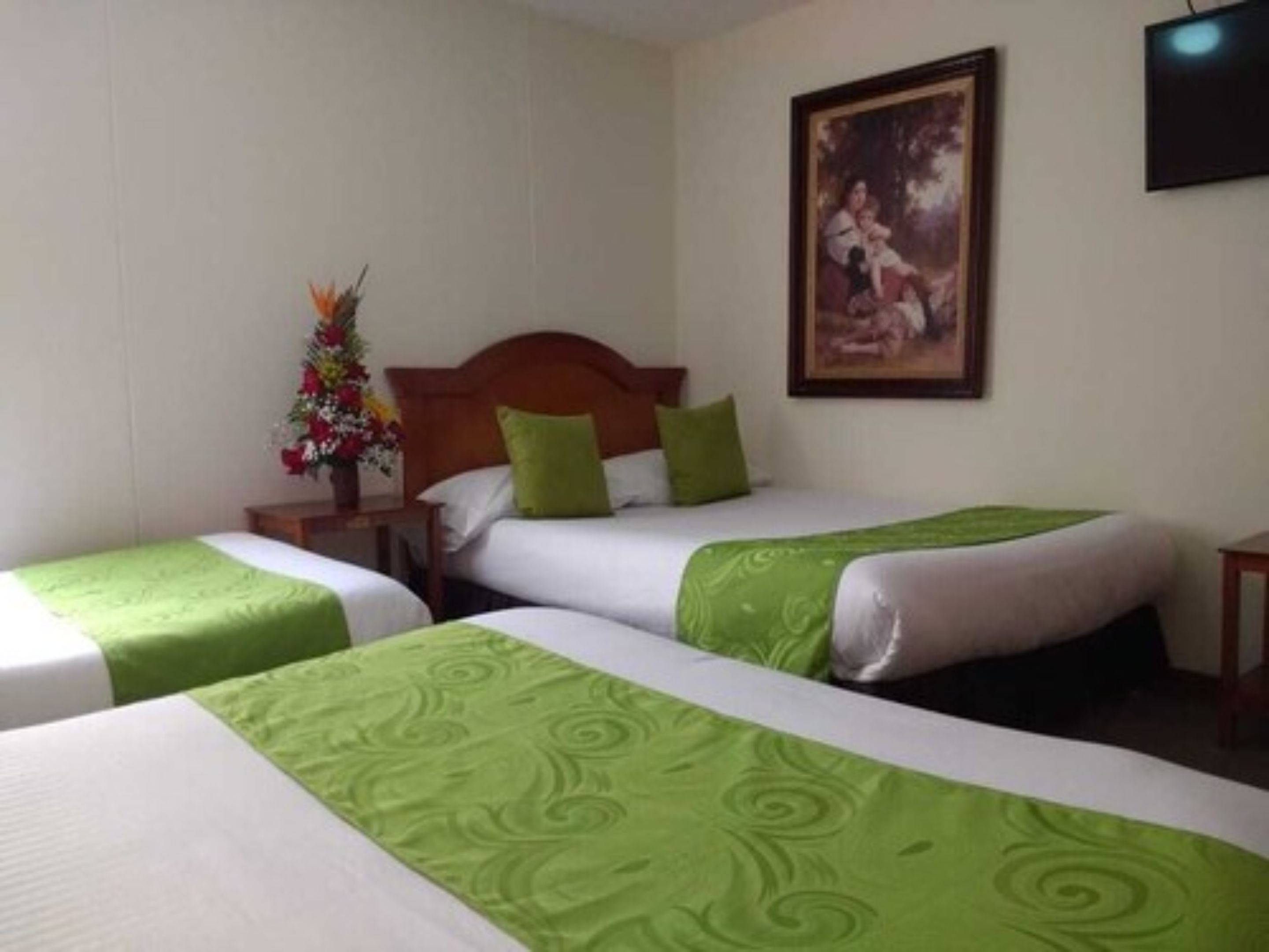 A hotel room with two single beds, each with a green and white bedspread, a wooden headboard, and a nightstand with a vase of flowers.