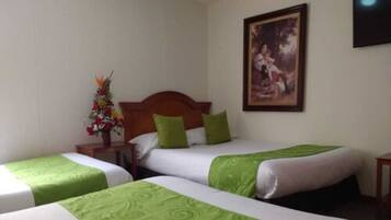 A hotel room with two single beds, each with a green and white bedspread, a wooden headboard, and a nightstand with a vase of flowers.