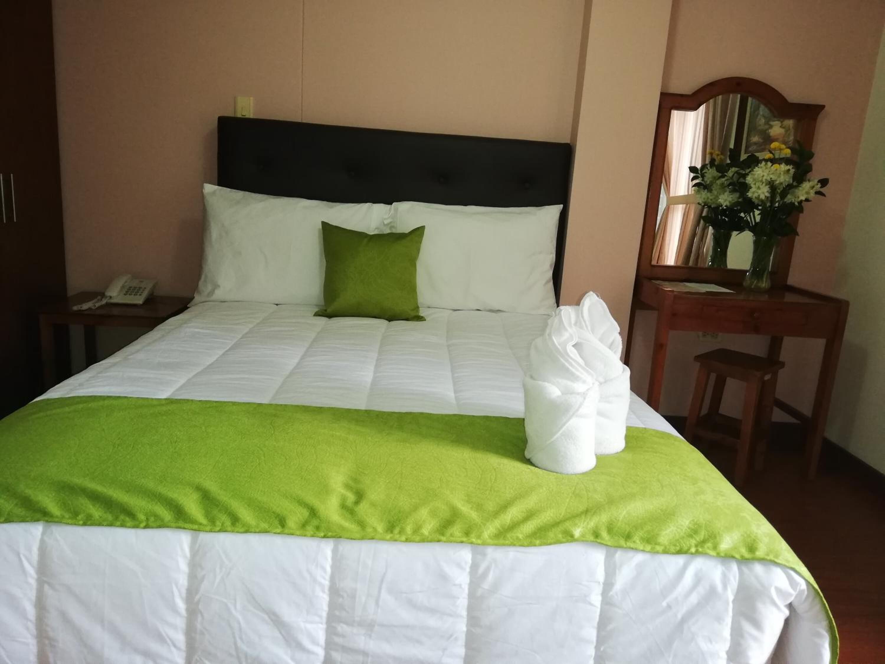 Una cama bien hecha con una colcha verde y blanca, una almohada verde y una toalla blanca sobre la cama. Hay una mesita de noche de madera con un teléfono y un espejo en la pared.