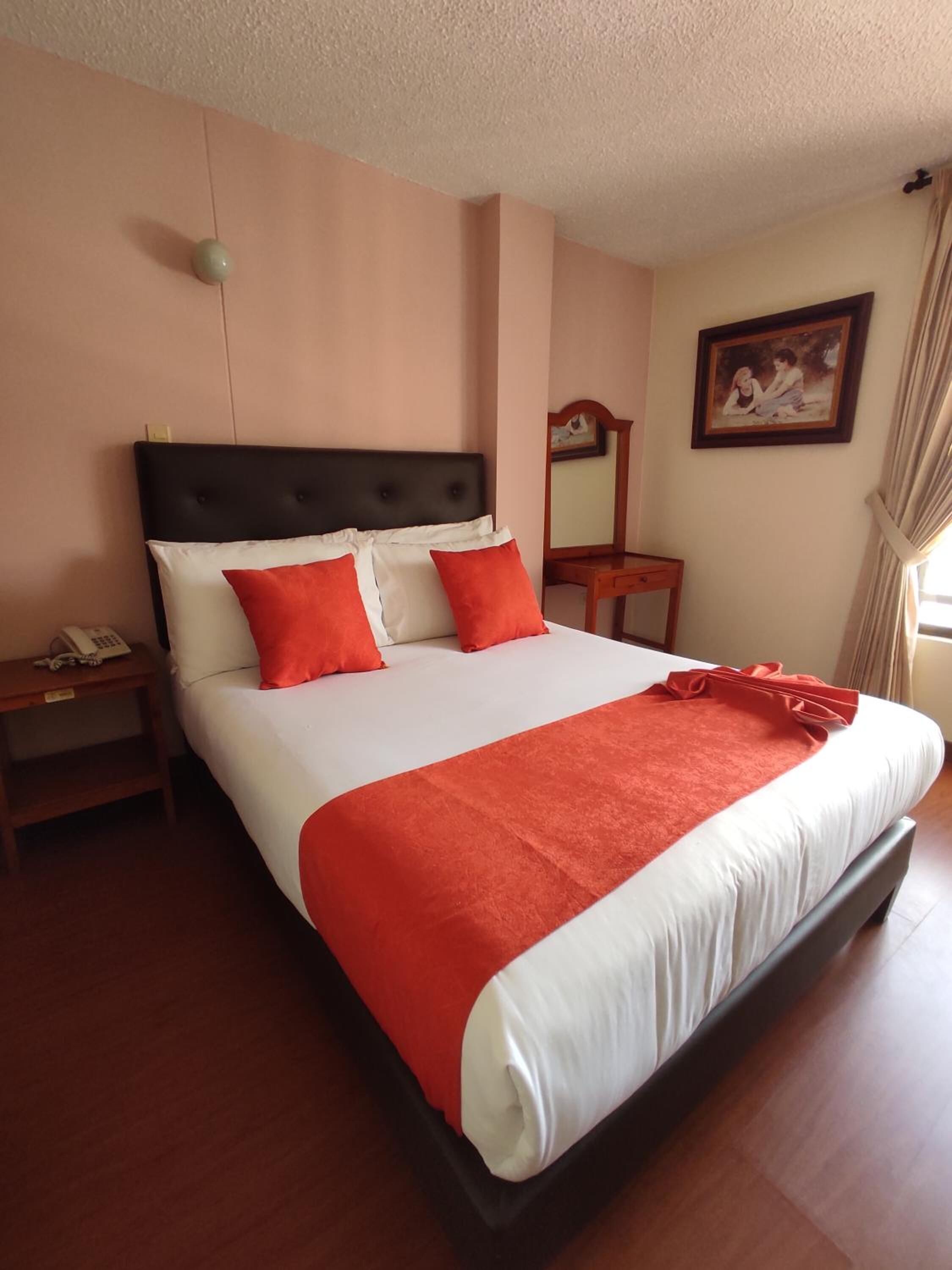 A neatly made bed with a white and orange bedspread, a wooden nightstand with a phone, and a mirror on the wall.