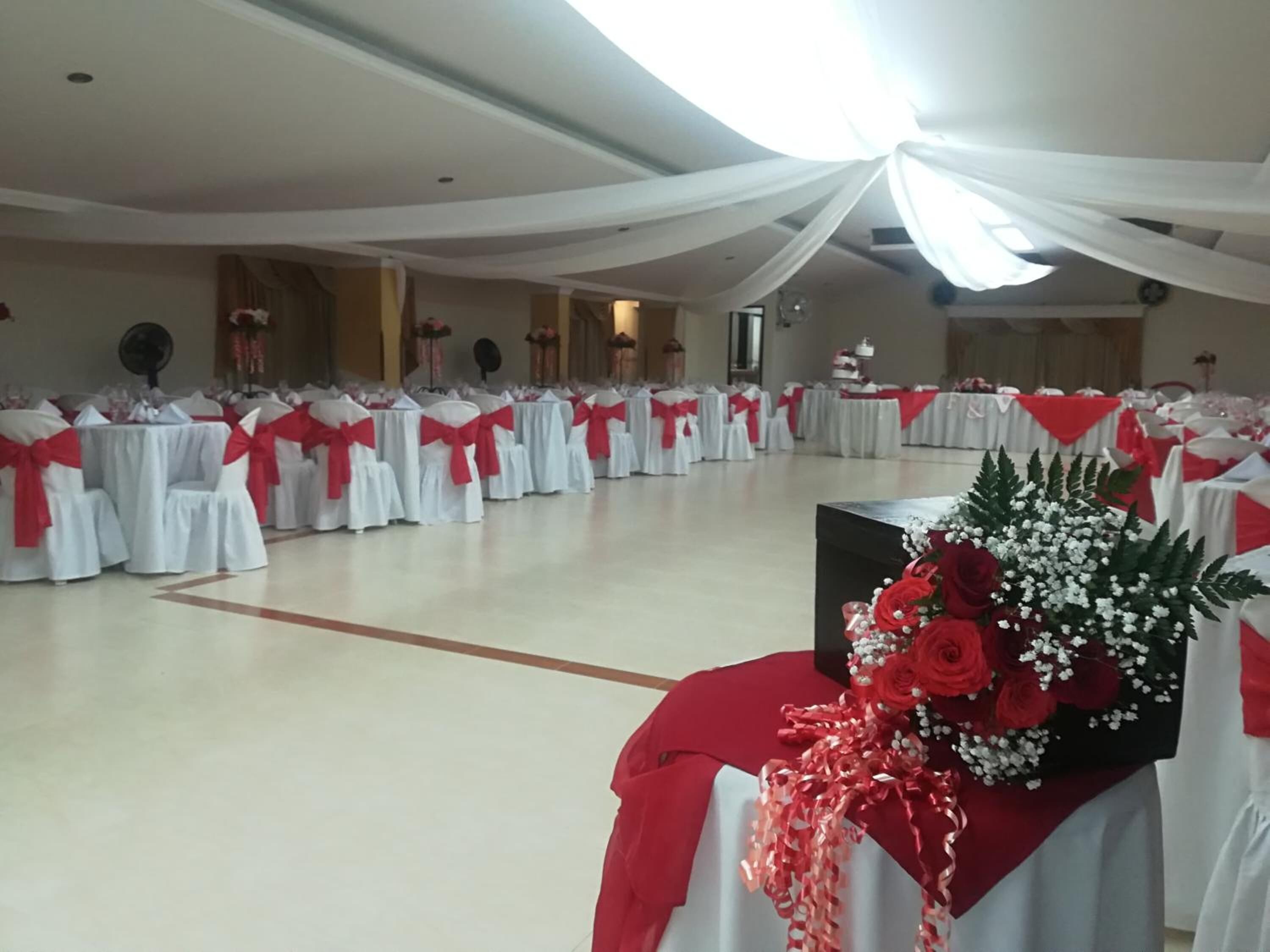 Um salão amplo com fileiras de mesas cobertas por toalhas brancas e fitas vermelhas, provavelmente montado para um evento.