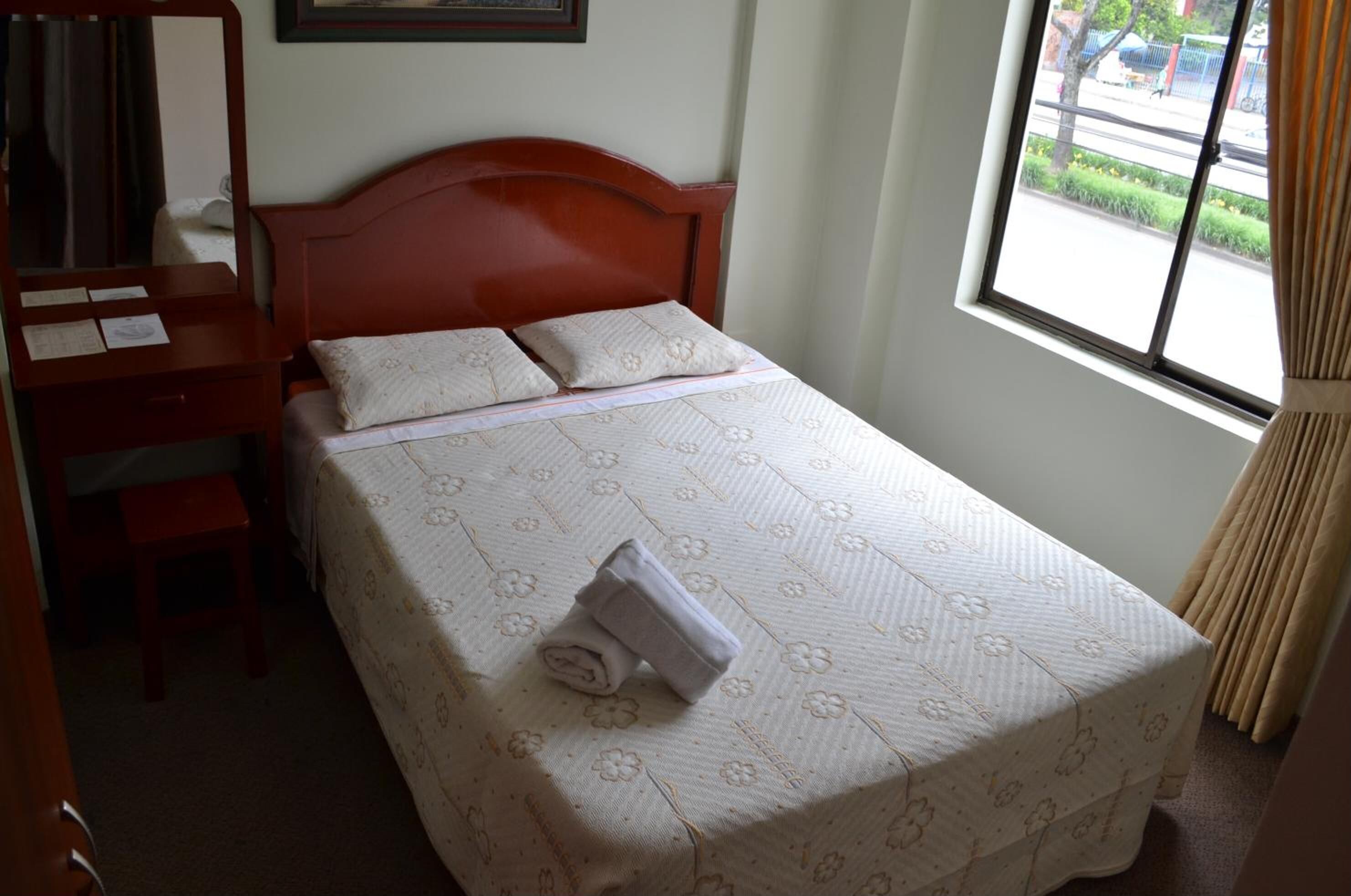 Una cama bien hecha con una colcha floral, una mesita de noche de madera con un espejo y una ventana con vistas a la calle.