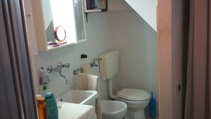 A bathroom with a sink, toilet, and mirror. There is a towel hanging on the wall and a shelf above the mirror. - Open space apartment (Cecina)
