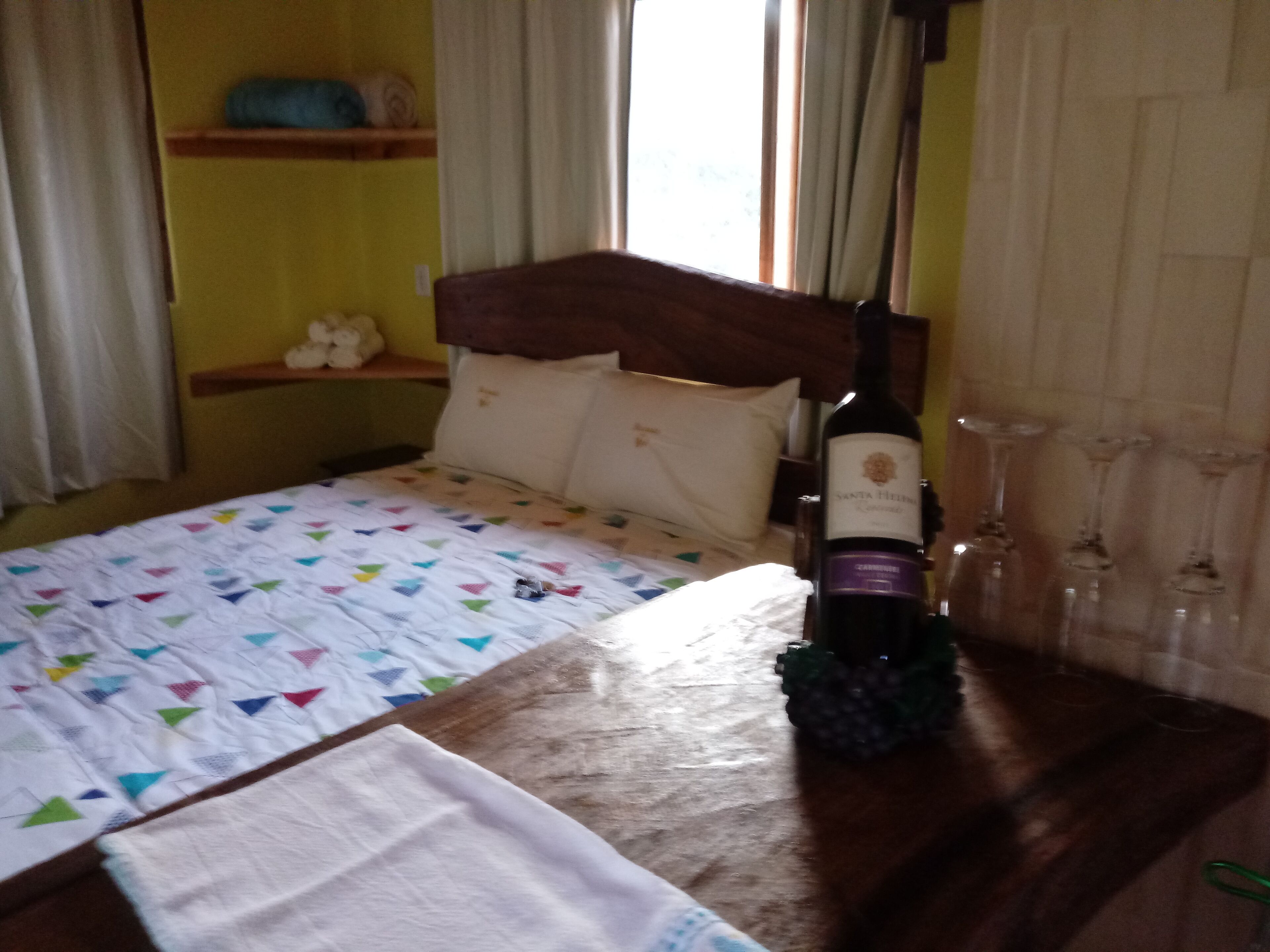 A bed with a colorful quilt, a bottle of wine, and wine glasses on a wooden table.