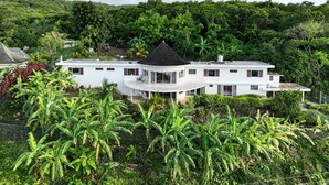 Een wit, twee verdiepingen hoog huis met een gebogen balkon, omringd door weelderige begroeiing en bananenbomen.