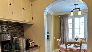 Una cocina con armarios blancos, salpicadero de ladrillo y un comedor con mantel floral y sillas de madera.