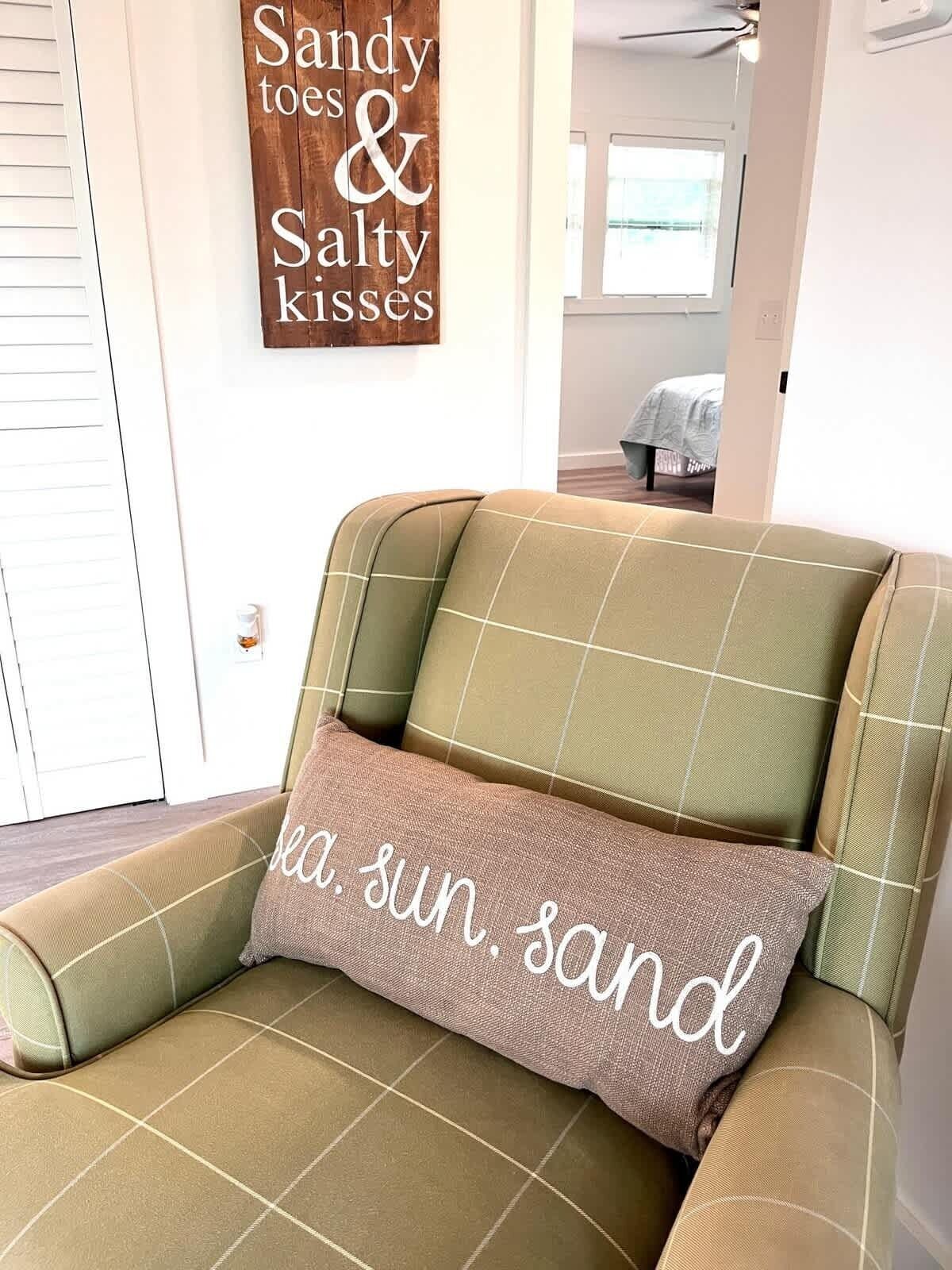 Ein Zimmer mit einem grünen Sofa, einem Kissen mit dem Aufdruck 'Meer, Sonne, Strand' und einem Schild an der Wand mit der Aufschrift 'Sandige Zehen und Salzküsse'.