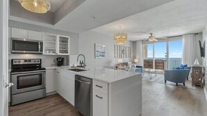 A modern kitchen with white cabinetry, a built-in microwave, and a dishwasher. There is a dining area with a blue sofa and a view of the outdoors. - Full Ocean Views, Steps to the Sand, Beach Chairs, Heated Pool/Hot Tub, Sleeps 8 (Miramar Beach)