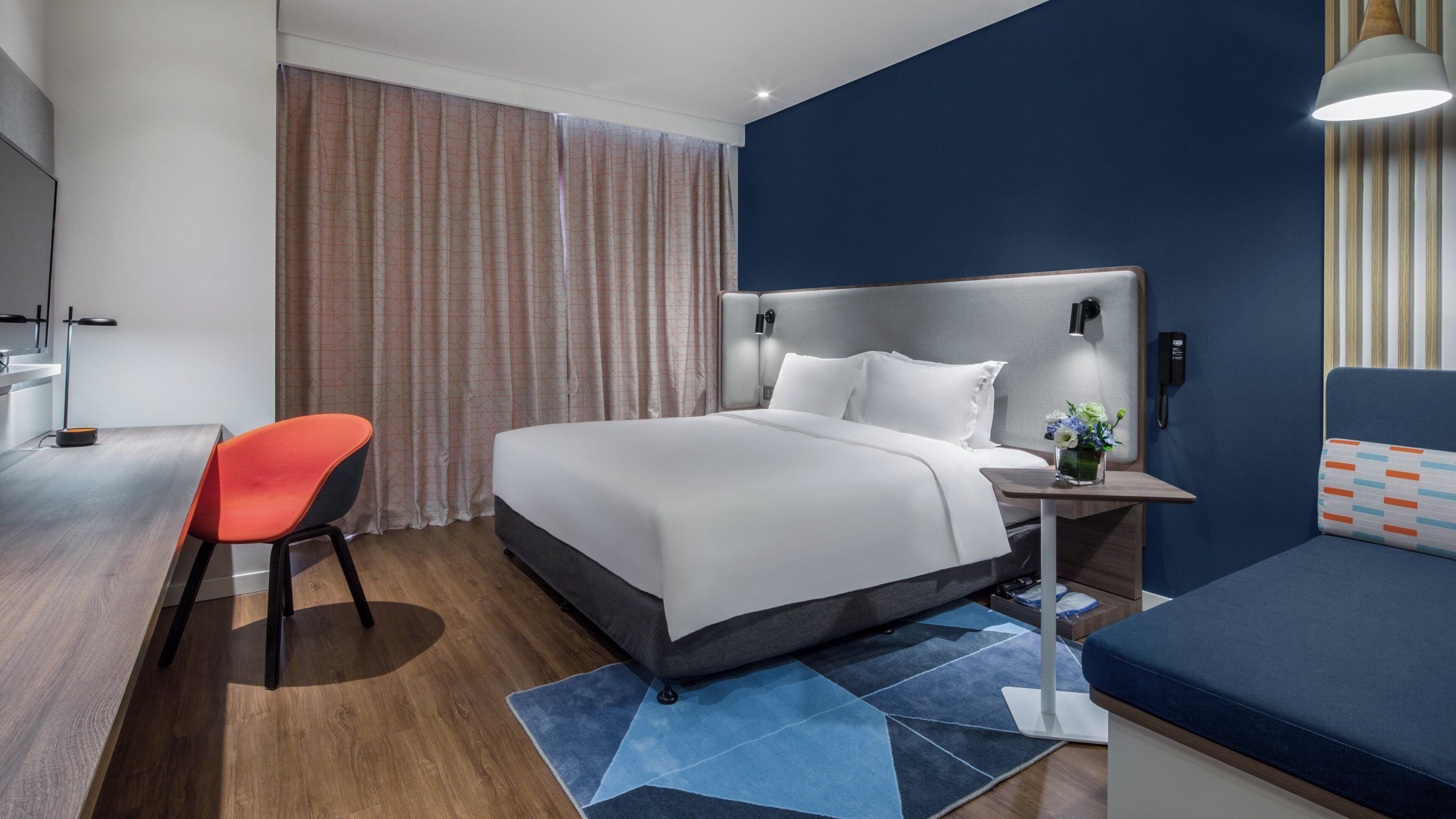 Quarto de hotel moderno com cama, escrivaninha, cadeira e mesa de cabeceira com abajur.