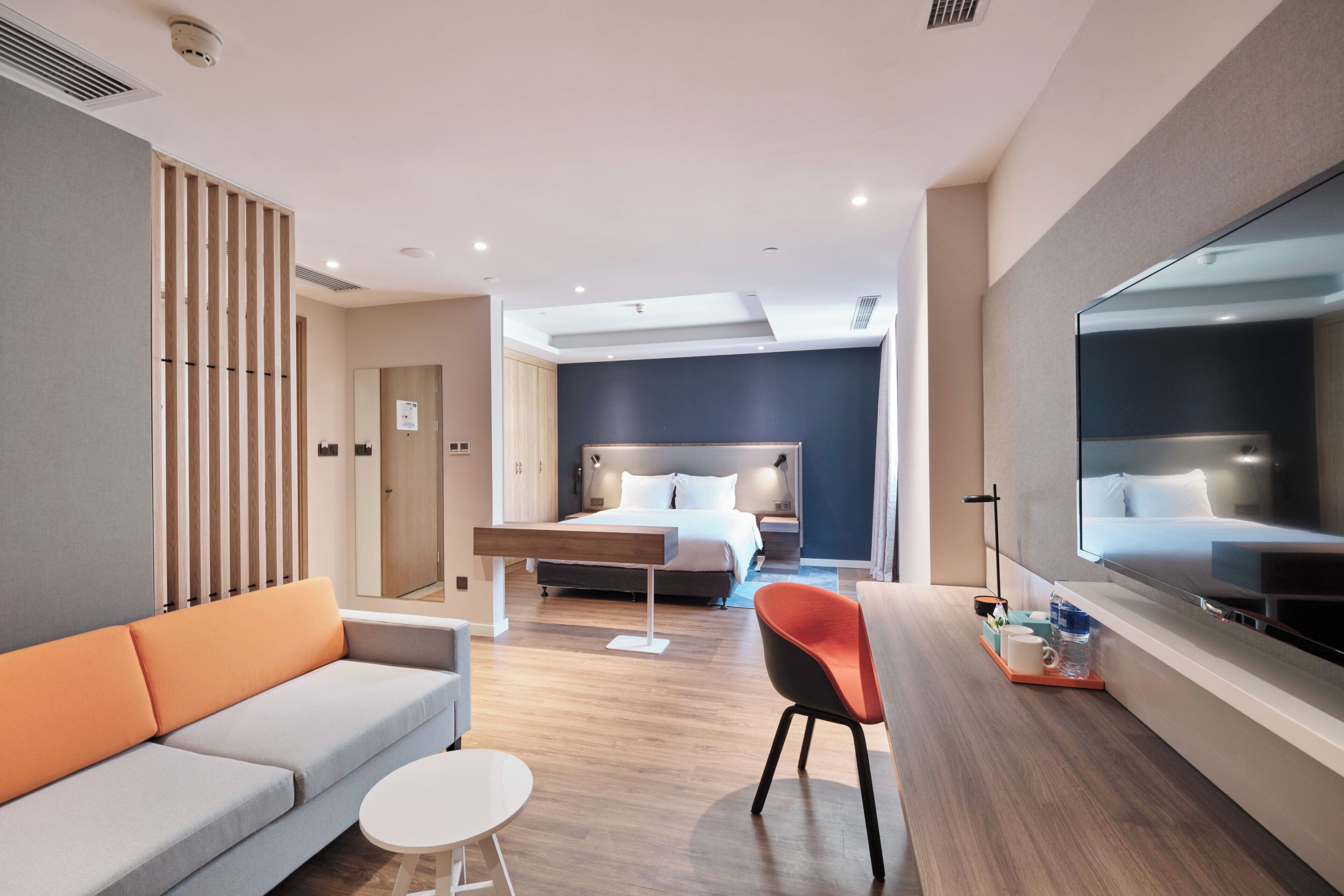 A modern hotel room with a sofa, a small round table, a bed with a headboard, a desk with a chair, a large mirror, and a television.