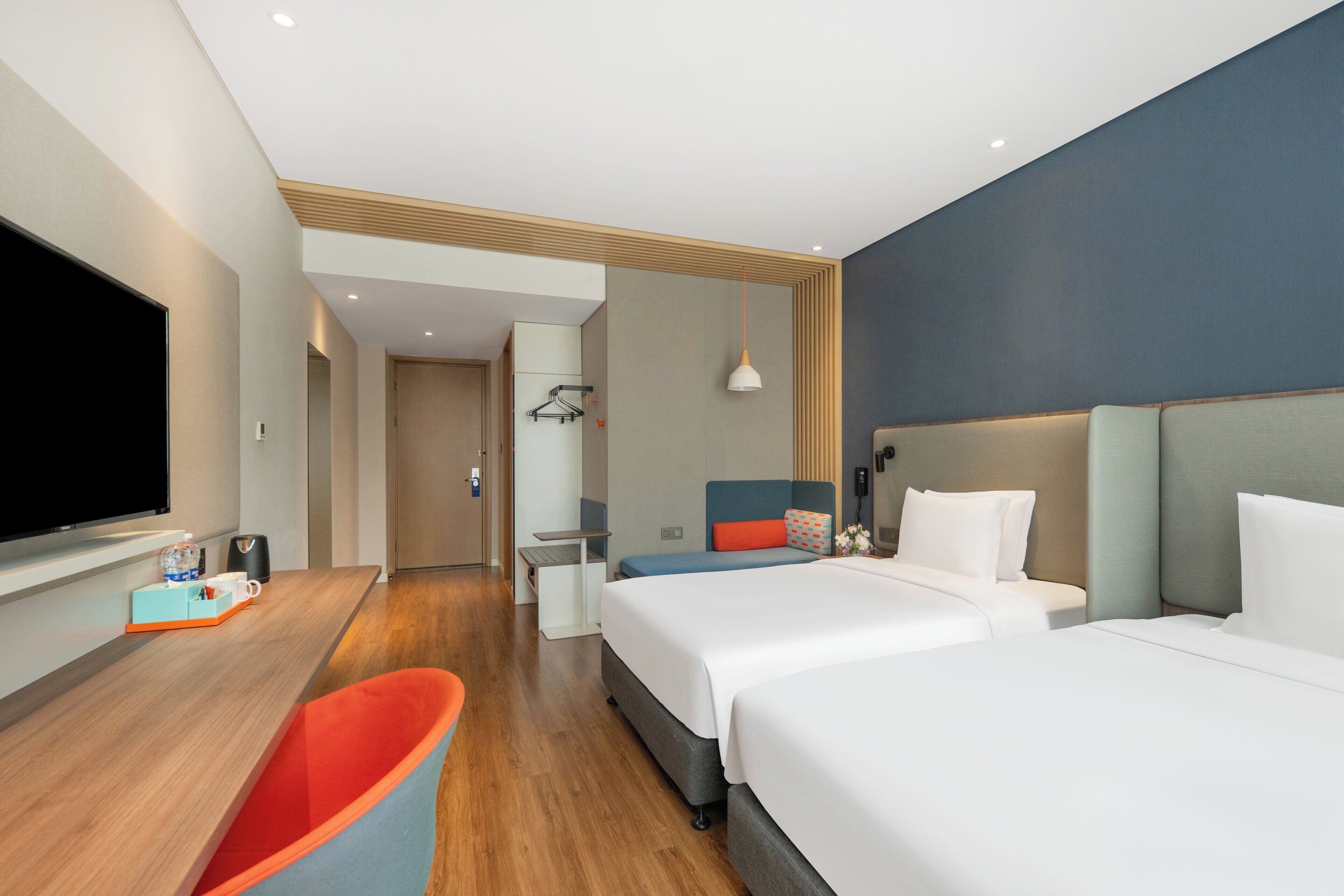 A modern hotel room with two beds, a flat-screen TV, a desk with a chair, and a bathroom visible through an open door.
