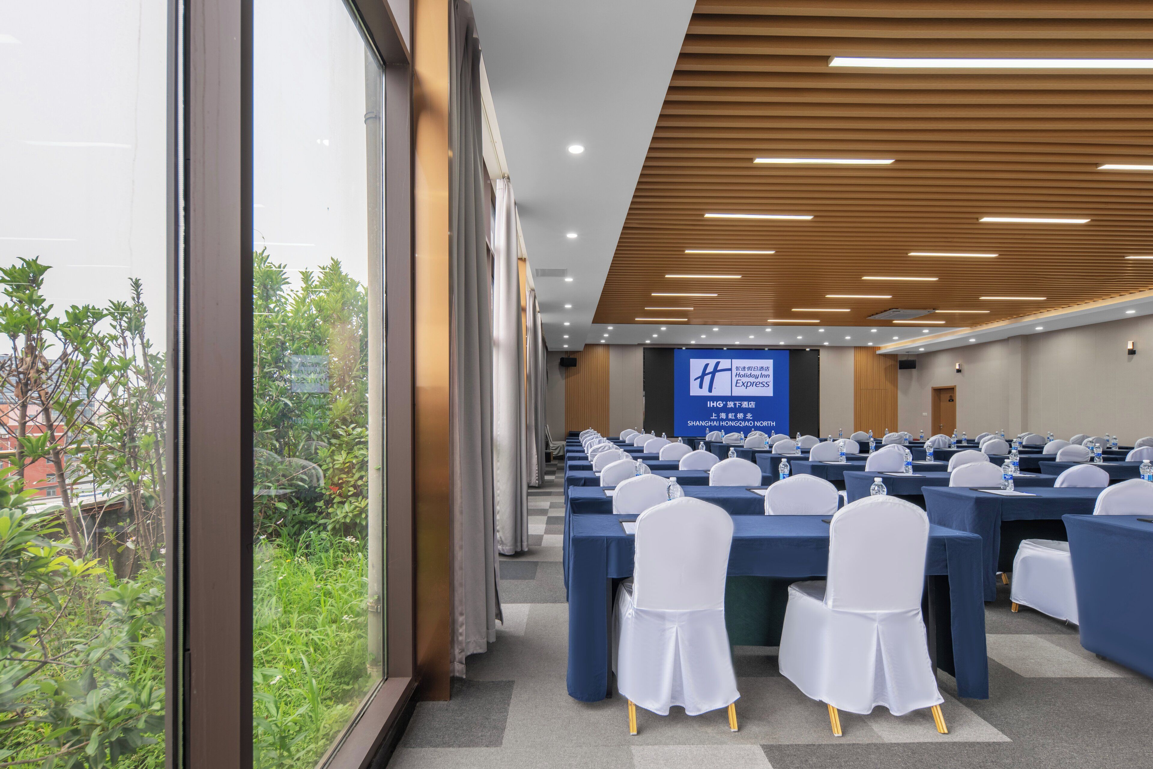 Un salón de conferencias con mesas y sillas dispuestas en filas, ventanas amplias y una pantalla de proyección.