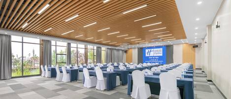 Un salón de conferencias con mesas largas, sillas blancas y una pantalla grande.