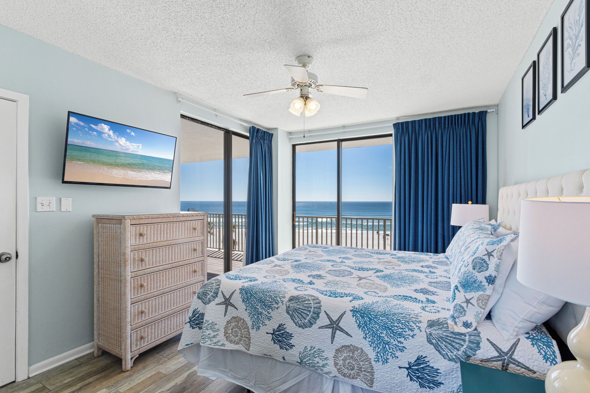 A bedroom with a bed, a dresser, a ceiling fan, a TV, and a view of the ocean.