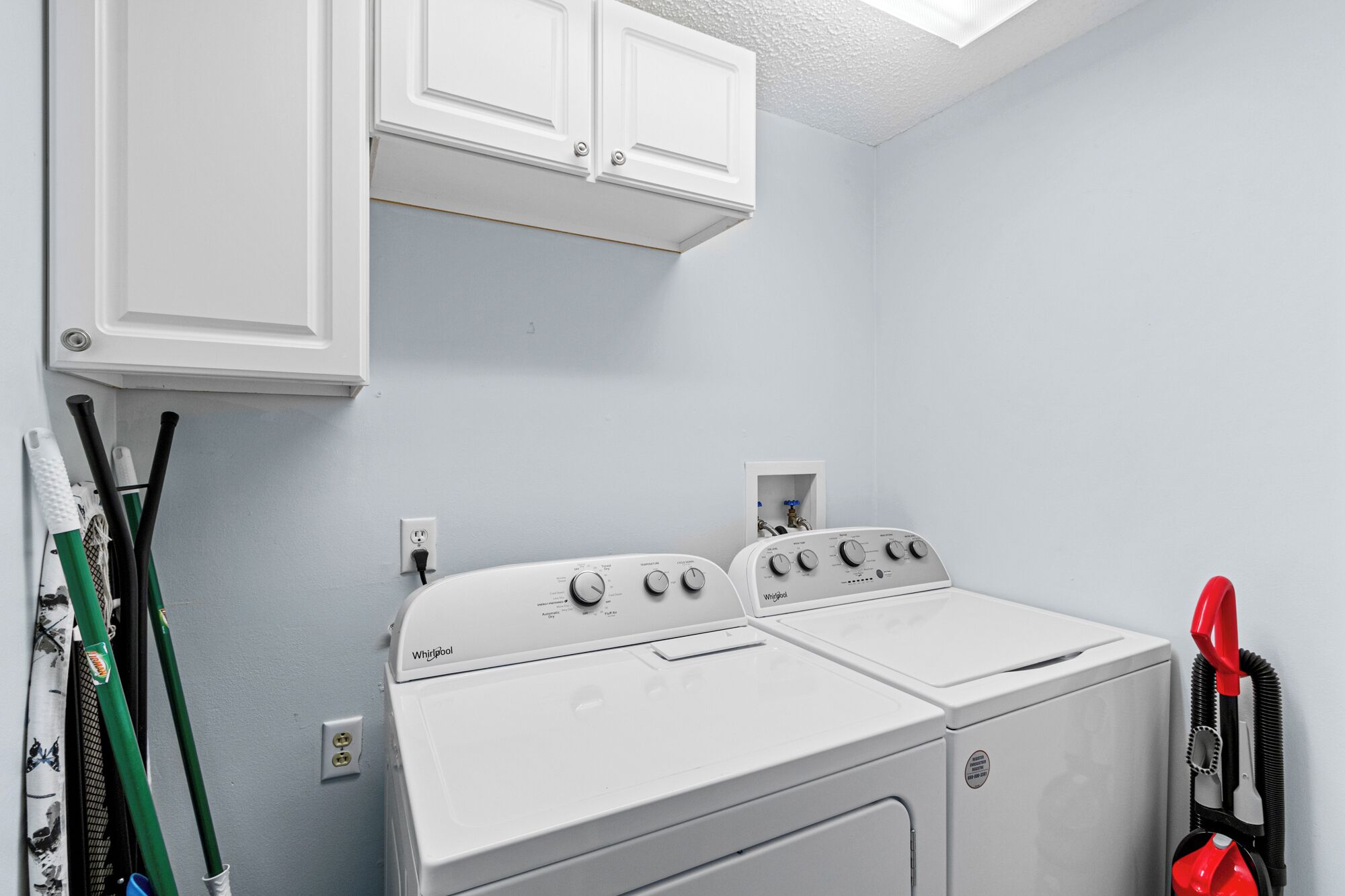 A laundry room with a front-loading washer and dryer, a white cabinet above, and a hose on the right.