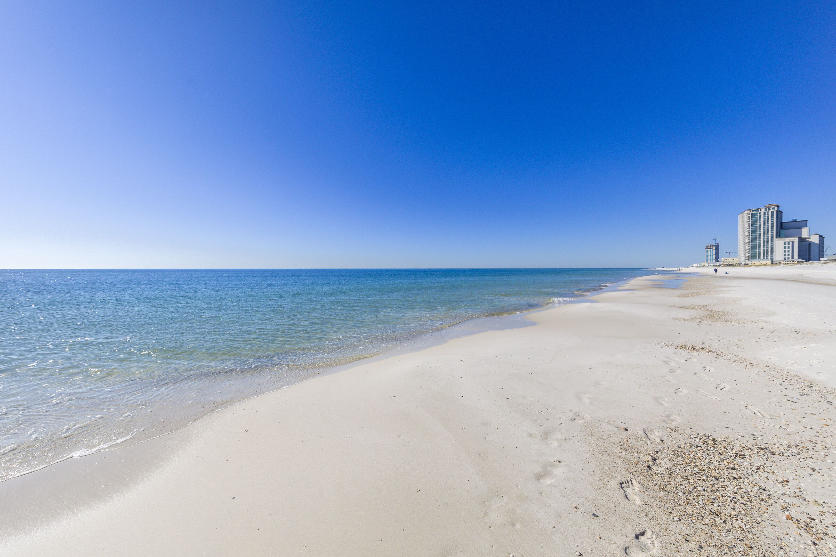 A beach with clear blue water and a sandy shore. There are buildings in the background.