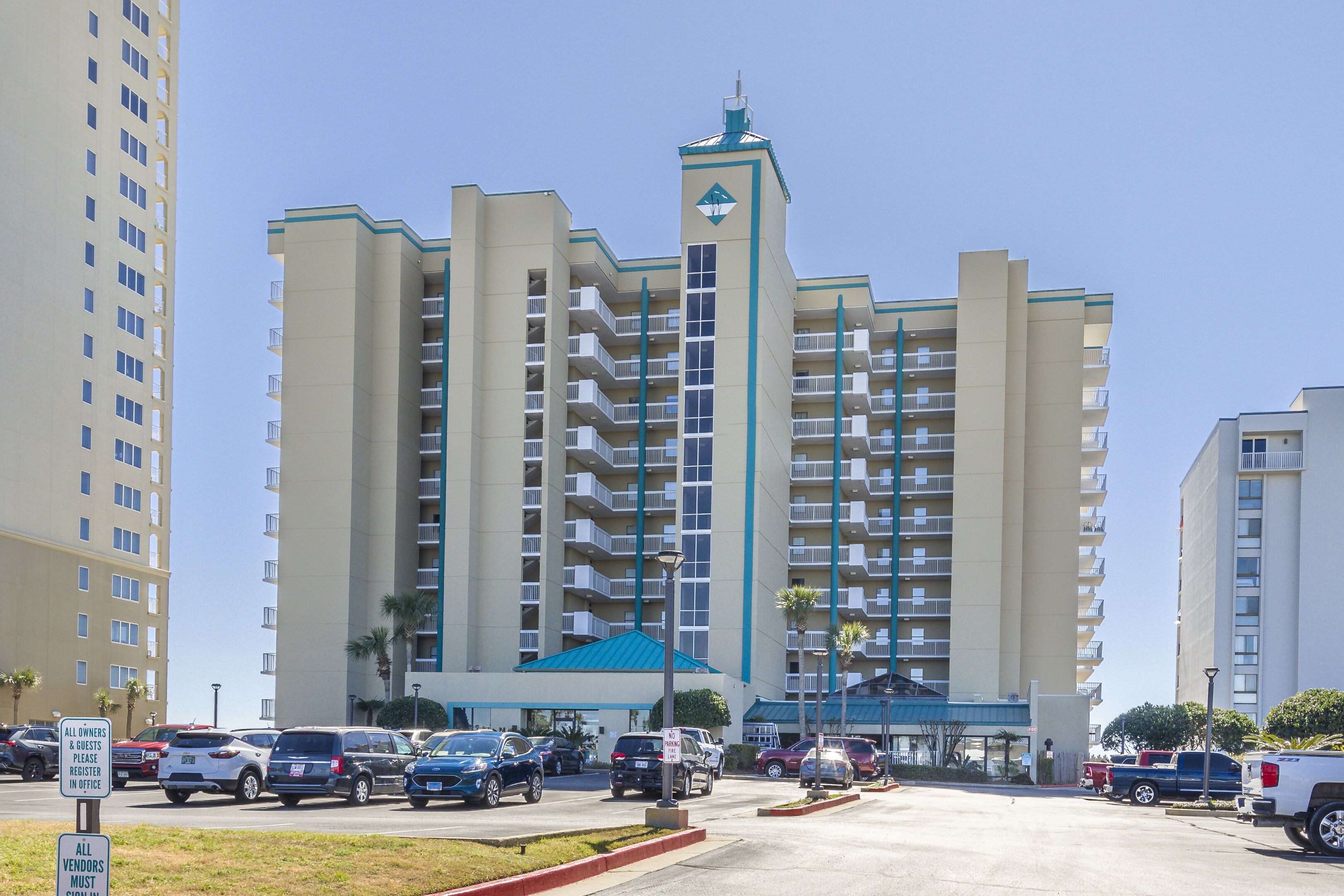 Un hôtel à plusieurs étages, doté d’une tour centrale, d’un parking contenant plusieurs voitures et d’un panneau indiquant qu’il est obligatoire pour les fournisseurs de s’inscrire.