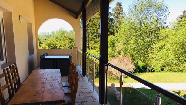 A covered outdoor dining area with stone flooring, wooden tables and chairs, and a view of a lush garden.