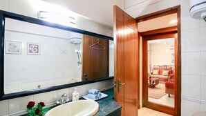Un baño con un espejo grande, un lavamanos y una ducha. Hay una puerta de madera que da a una sala de estar con un sofá y una mesa de centro.
