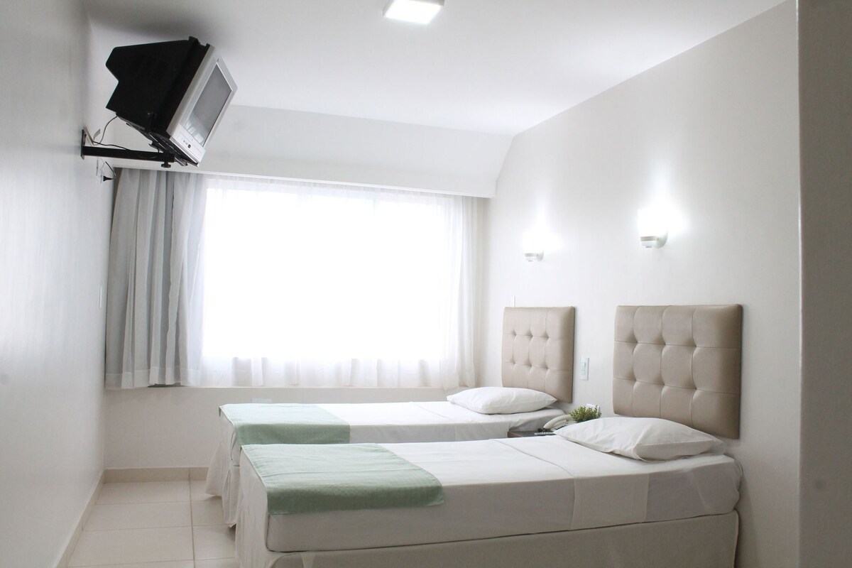 A hotel room with two beds, a flat-screen TV mounted on the wall, a window with white curtains, and a wall-mounted light fixture.