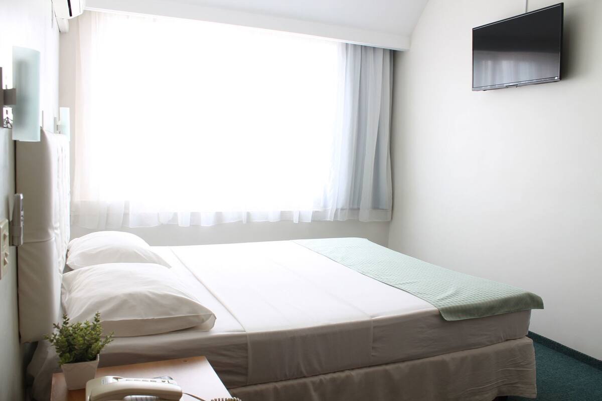 Een hotelkamer met een bed, nachtkastje met telefoon, kleine plant, raam met dunne gordijnen en een wandtelevisie.