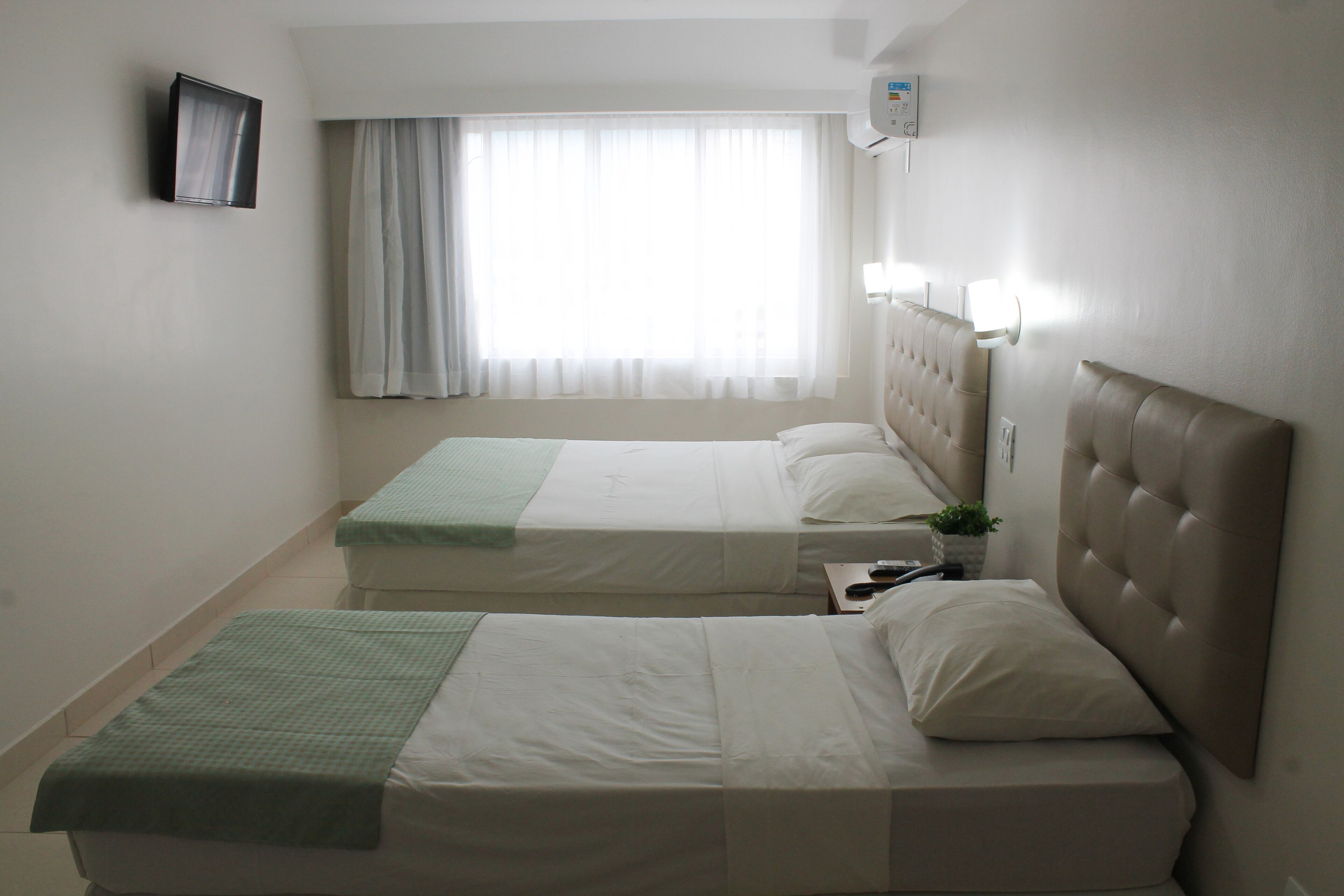 A hotel room with two beds, a television, a window with curtains, and a wall-mounted air conditioner.