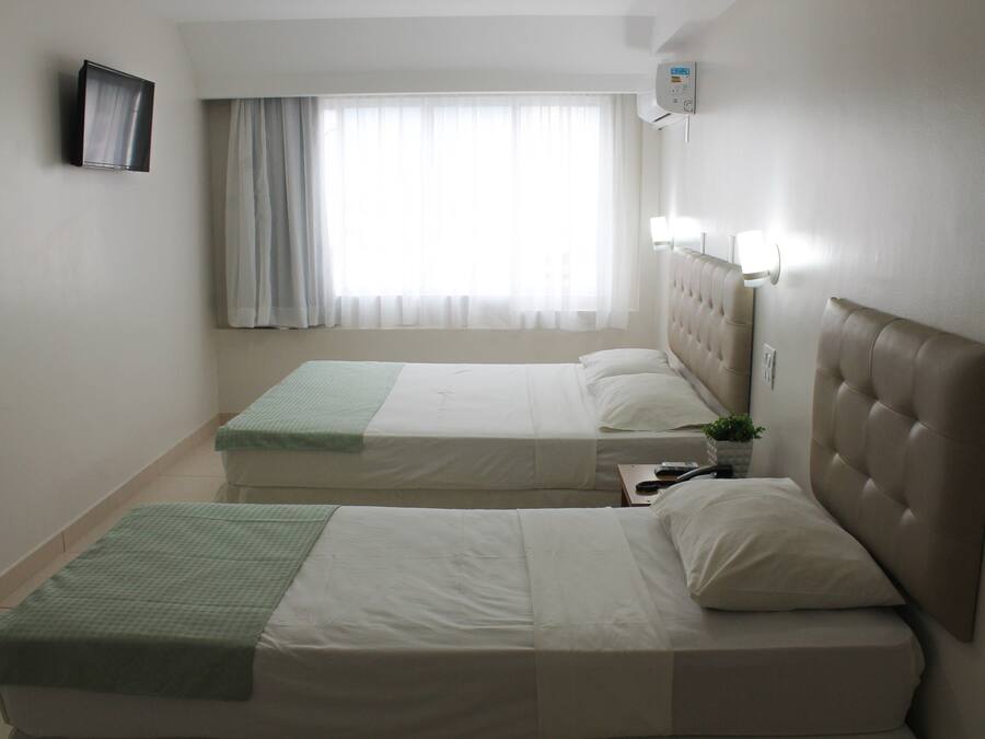 A hotel room with two beds, a television, a window with curtains, and a wall-mounted air conditioner.