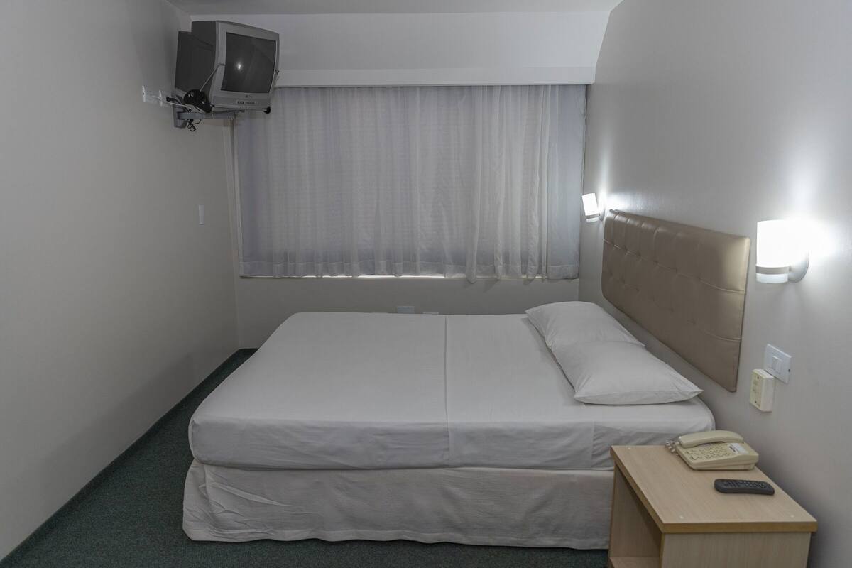Een hotelkamer met een bed, een aan de muur bevestigde televisie, een nachtkastje met telefoon en afstandsbediening, en een raam met gordijnen.