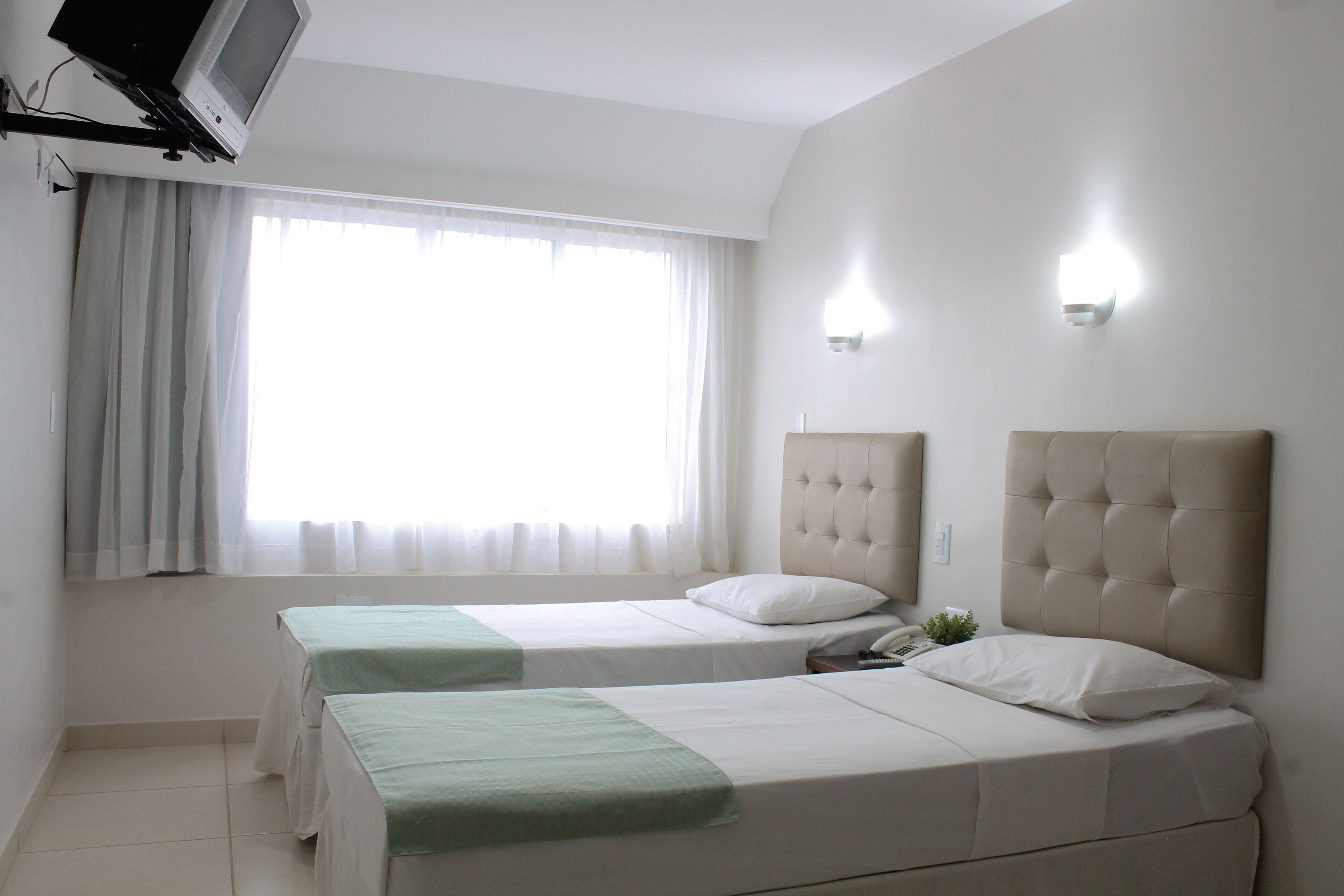 A hotel room with two beds, a television, a window with curtains, and a wall-mounted light.