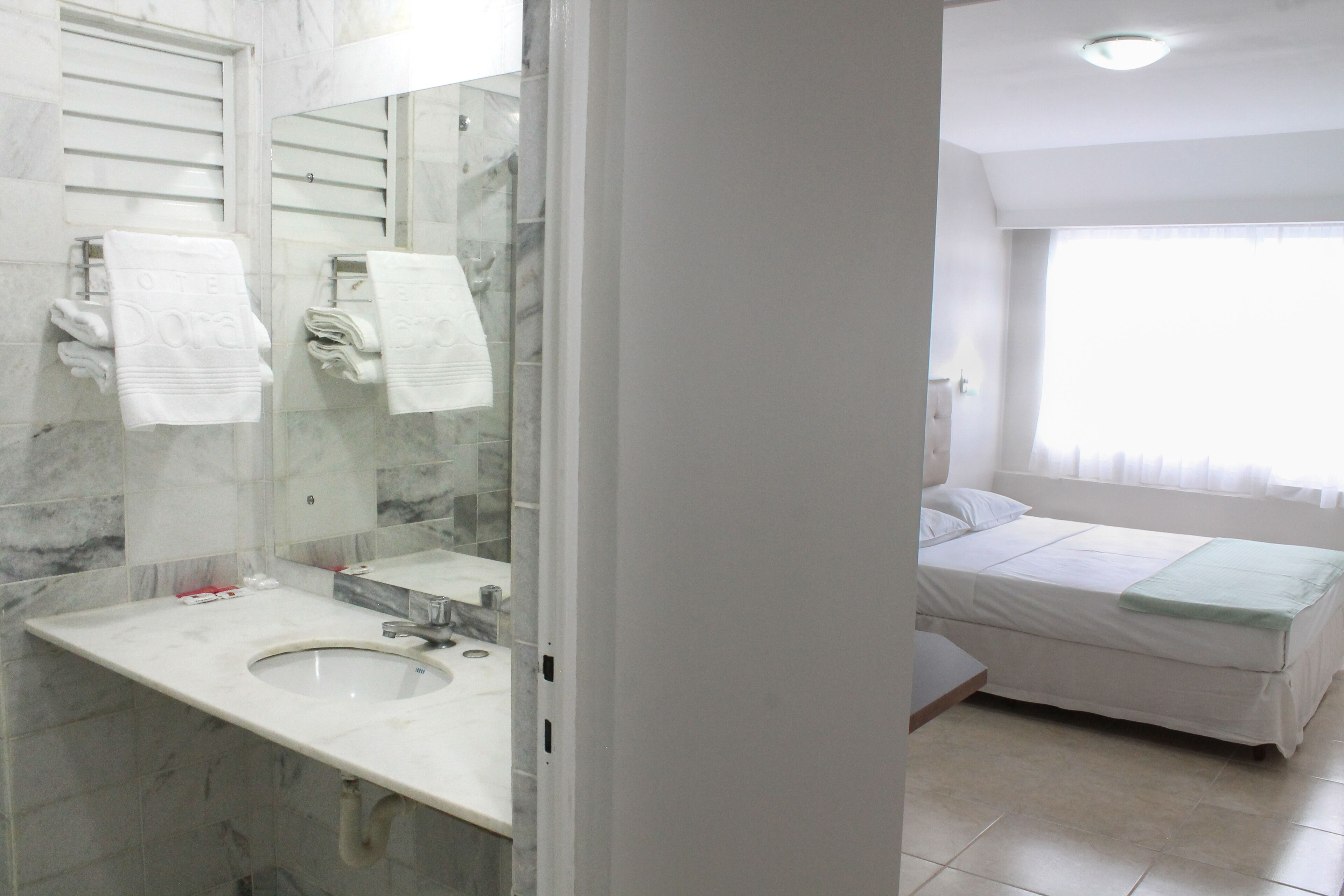 A bathroom with a marble sink, a mirror, and a bed with a green blanket in the adjacent room.