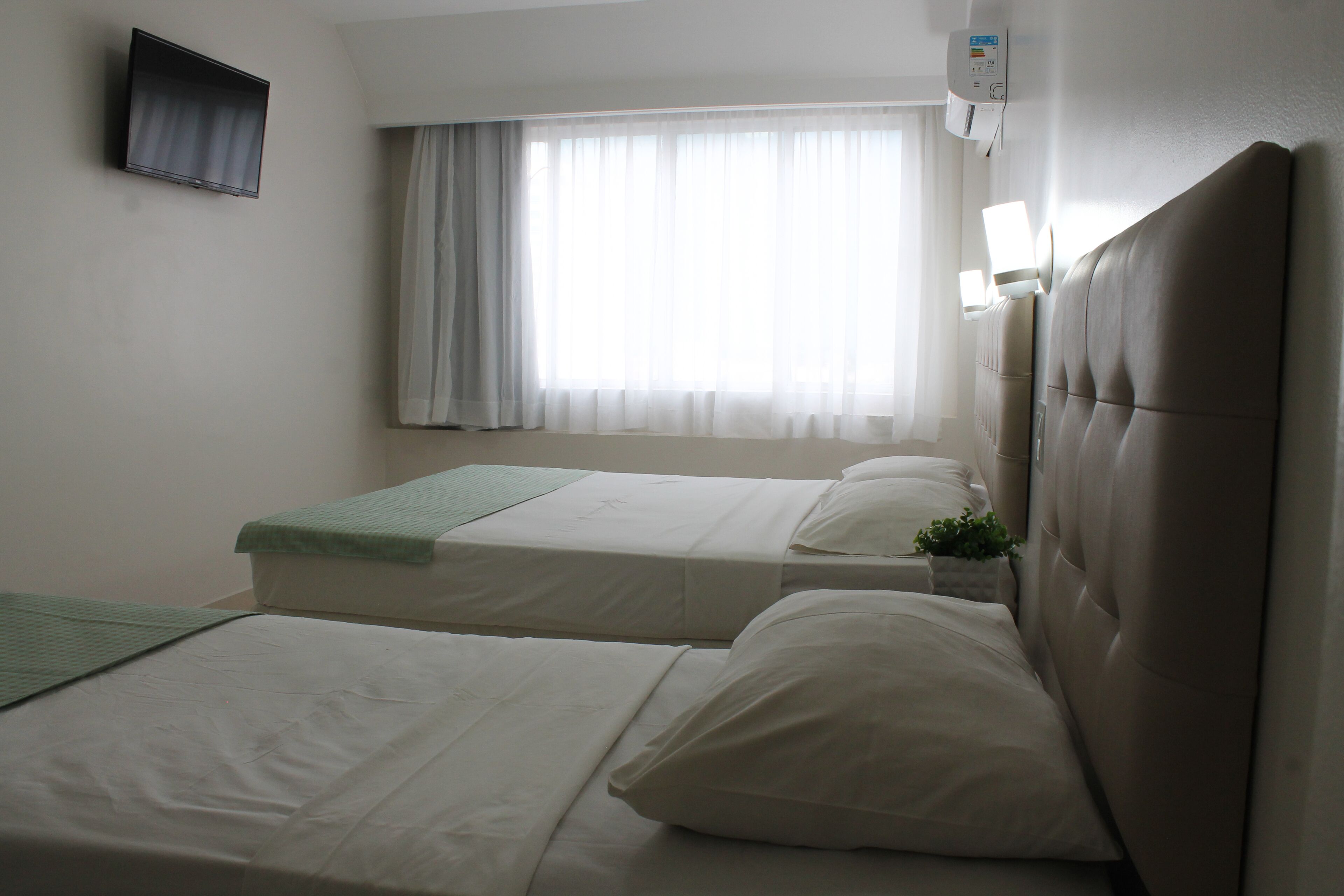 A hotel room with two beds, a flat-screen TV mounted on the wall, a window with sheer curtains, and a wall-mounted air conditioning unit.