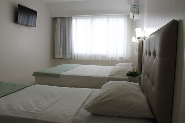Habitación de hotel con dos camas, un televisor de pantalla plana montado en la pared, una ventana con cortinas traslúcidas y un aire acondicionado también fijado a la pared.