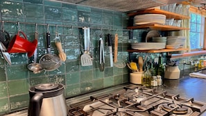 A kitchen with a gas stove, hanging utensils, and a shelf with plates and bottles.