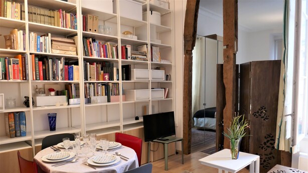 A dining area with a table set for two, a bookshelf filled with books, and a television on a stand. - Appart central near Louvre  (Paris)
