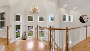 A spacious interior with a high ceiling, a wooden staircase, and multiple windows offering views of the outdoors. - Sound Escape: Elevator, Hot Tub, Private Pool, Soundfront Location (Corolla)