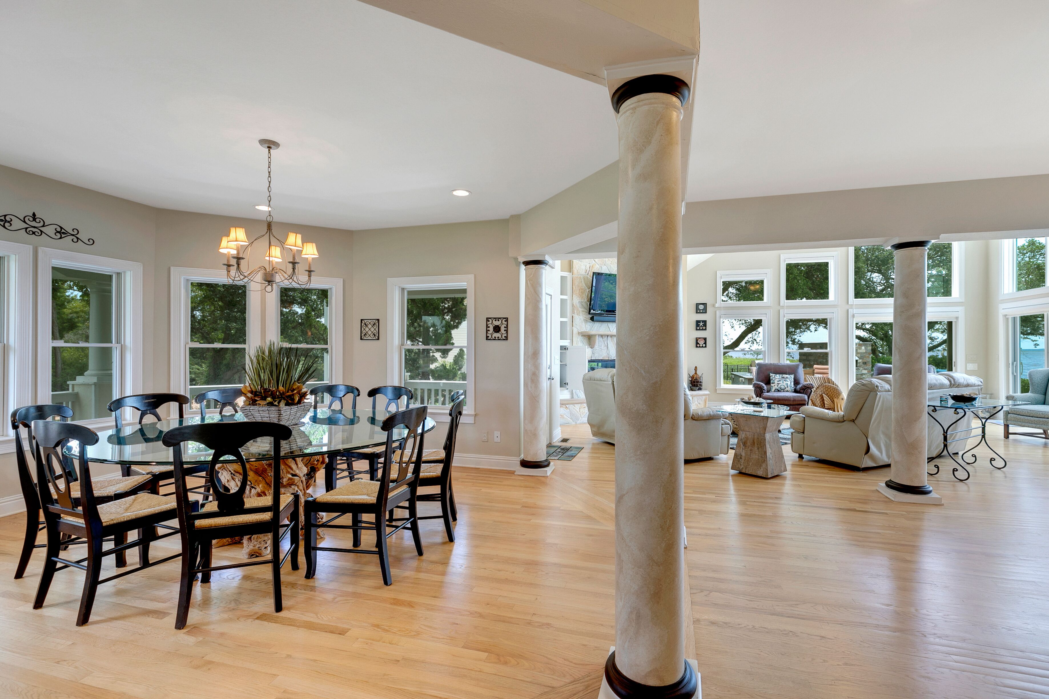 A spacious living room with a large glass dining table surrounded by chairs, a chandelier, and large windows offering a view of the outdoors.