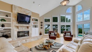 A spacious living room with a stone fireplace, large windows, and a view of the outdoors. - Sound Escape: Elevator, Hot Tub, Private Pool, Soundfront Location (Corolla)