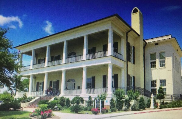 A two-story building with a balcony on the ground floor, surrounded by greenery.