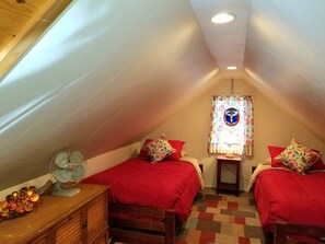 Ein kleines, gemütliches Schlafzimmer im Dachgeschoss mit zwei Betten, einer Holzkommode, einem Ventilator und einem Buntglasfenster.