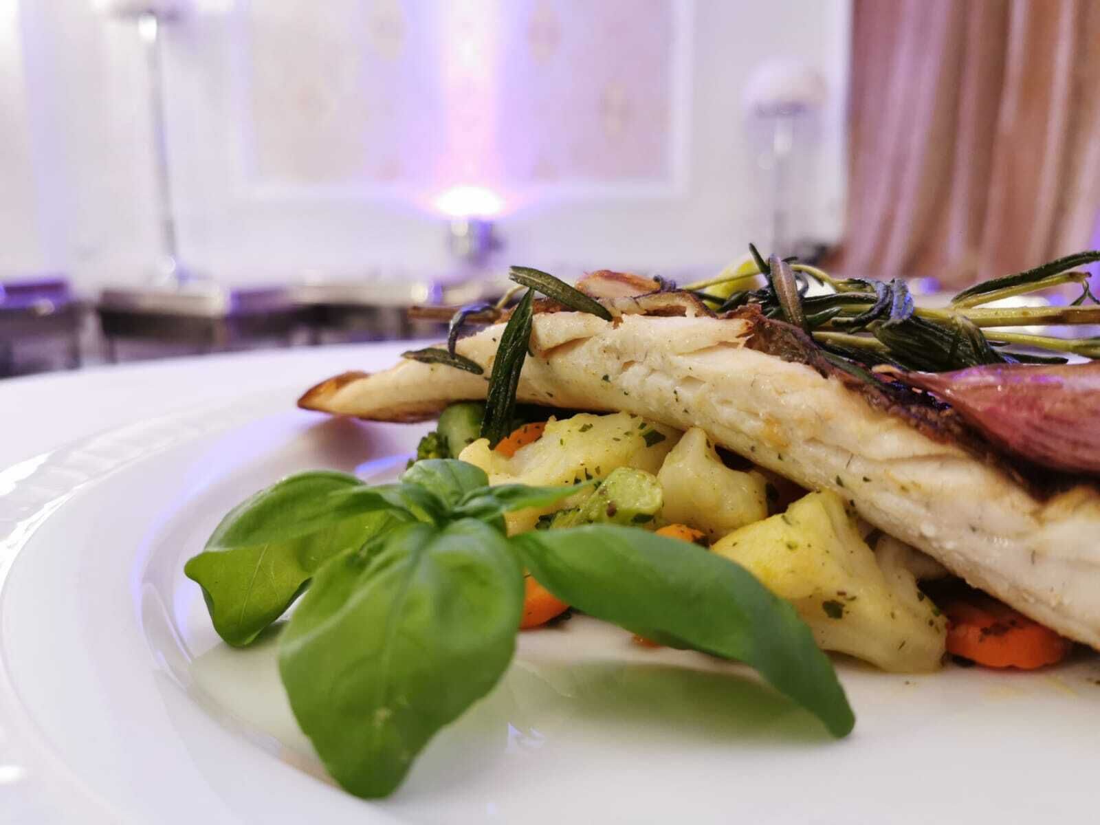 A plate of food with a white plate, a piece of fish, roasted vegetables, and a basil leaf.